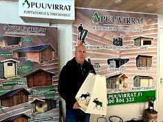 Kimmo Hynynen kantaa käsissään komeaa hirvikuvioista puunkantotelinettä. Tuotetta on eri aiheilla ja väreillä. Taustalla näkyy valikoima myynnissä olevista liitereistä, laavuista ja vajoista sekä roskiskatoksista.