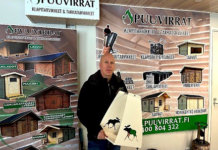 Kimmo Hynynen kantaa käsissään komeaa hirvikuvioista puunkantotelinettä. Tuotetta on eri aiheilla ja väreillä. Taustalla näkyy valikoima myynnissä olevista liitereistä, laavuista ja vajoista sekä roskiskatoksista.